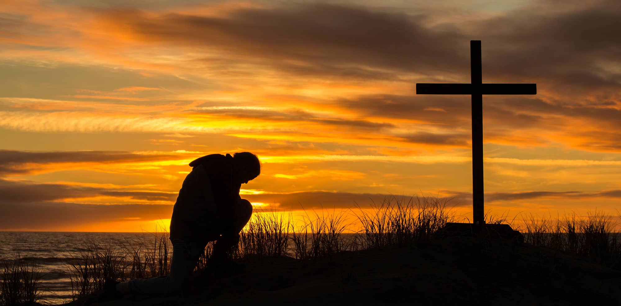 Man praying in front of cross at sunset